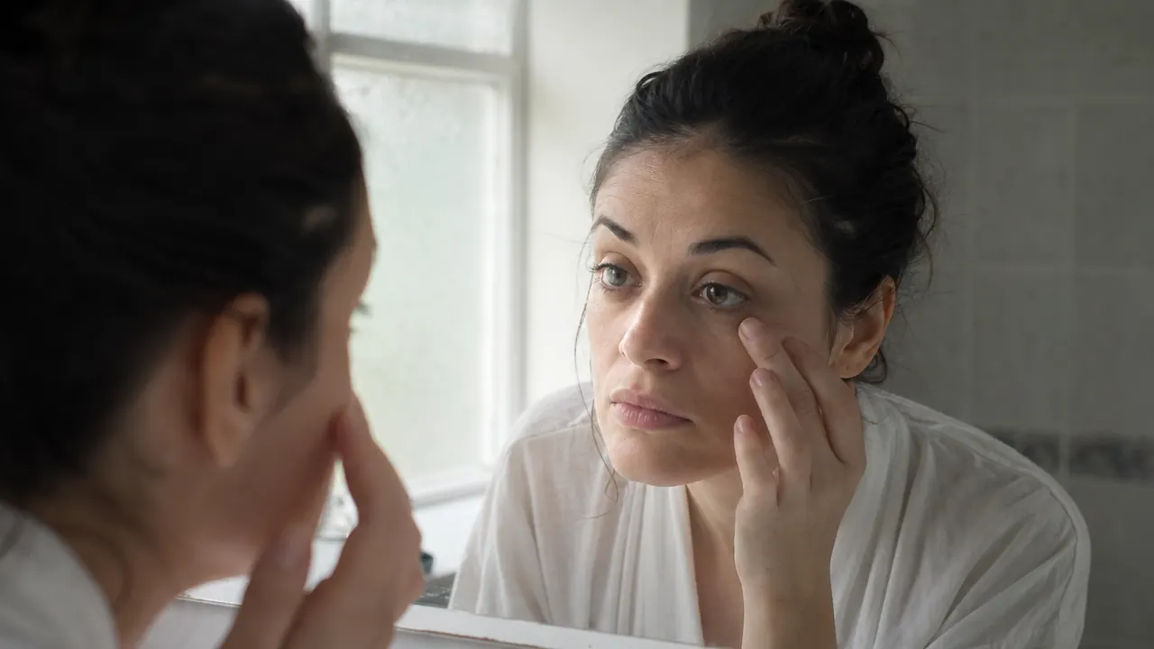 Person looking in a bathroom mirror examining the skin under their eyes in morning light